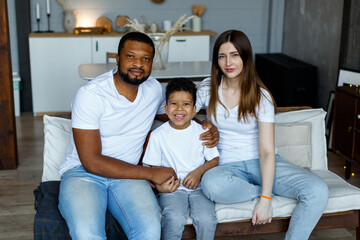 Loving black family relaxing at sofa in the morning, happy mixed race parents laughing cuddling having fun with cute little kid child son playing enjoying moments together. High quality photo