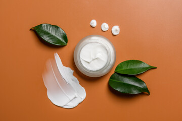 Glass skin cream jar on brown background with smear or swatch and leaves