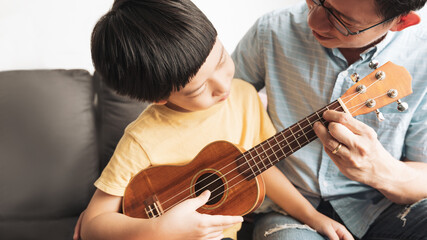 Asian father teaching his little boy to play ukulele with joy a small, guitar-like instrument in...