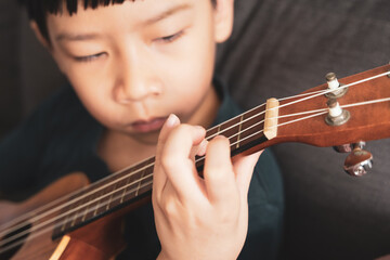 Young Asian kid playing ukulele and practicing his fingers position alone with calm and...
