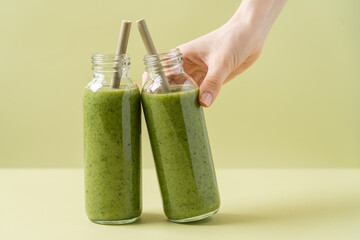 Green smoothie in glass bottles with bamboo tubes. Woman's hand holding bottle. Monochrome green background. Minimalistic concept advertising a product, clean, healthy food - Powered by Adobe