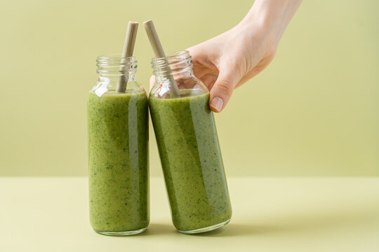 Green smoothie in glass bottles with bamboo tubes. Woman's hand holding bottle. Monochrome green background. Minimalistic concept advertising a product, clean, healthy food
