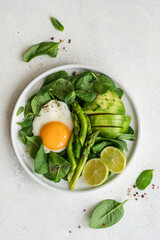 Healthy green breakfast with fried egg. Asparagus, spinach, avocado and egg in plate on white stone background