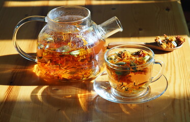 Herbal medicinal tea with dried flowers of marigolds and calendula. Glass teapot with a cup of tea in the evening light.