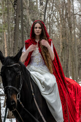 beautiful woman in white dress and red cape with black horse in spring forest 