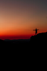 Fototapeta na wymiar Red sunset with mountain silhouette. A man on mountain in sunset. Traveler on rock waving hands.