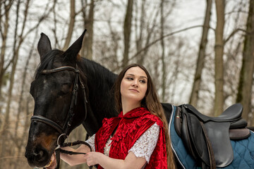 beautiful woman in white dress and red cape with black horse in spring forest 