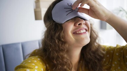 Attractive curly haired woman in funny sleeping mask with bunny face shoots video with phone dancing on bed in hotel room in sunny morning closeup