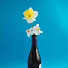 Tuinposter Yellow narcis and white spring flowers in a botlleof wine on a blue background,minimal composition © Mira