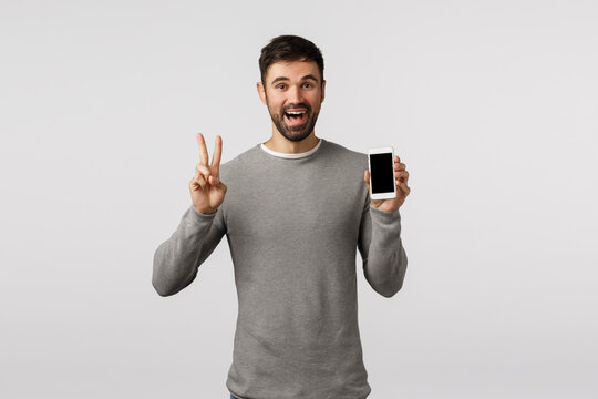 Friendly And Carefree Funny Bearded Gay Man In Grey Sweater Edit Profile Picture Online Dating App, Make Peace Sign, Showing Smartphone Display And Smiling Happily, Standing White Background