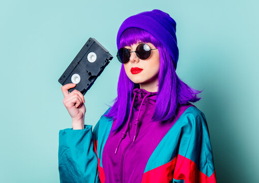 Stylish white girl with purple hair, 80s tracksuit and sunglasses hold video tape on blue background