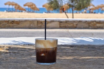 Closeup of an iced coffee with a straw in front of the mylopotas beach in Ios Greece