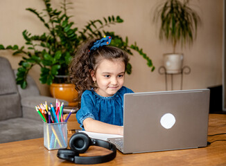 Happy little girl in headphones smiling and making notes in notepad while communicating with teacher through video chat app during online education at home 