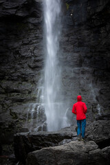 Man stands on rock with red jacket, looks at Fossa waterfall on Streymoy Island, Faroe Islands.