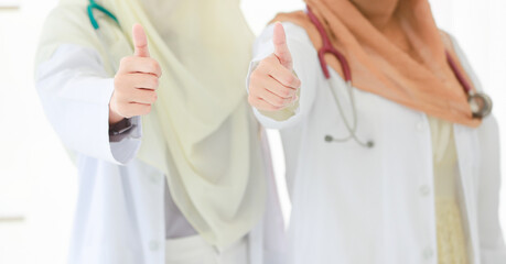Two adult Muslim doctors, wearing hijab with a stethoscope around neck looking camera, and showing a thumb up gesture with hands. Concept of successful treatment. Selective focus at the hands
