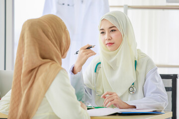 Horizontal portrait shot of attractive smiling adult Muslim woman wearing brown hijab sitting at the table, looking and concentrated talking to doctor. Selective focus at a patient with blurred doctor