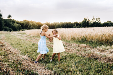 Naklejka na ściany i meble Family lifestyle and summertime concept. Two emotional sisters dance in the field with rye at sunset.