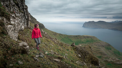Obraz premium Female hiker with magenta jacket and tights in mountain landscape of Streymoy island with cloudy sky, Faroe Islands.