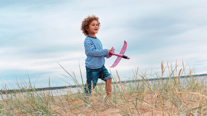 Naklejka na ściany i meble Small kid (boy, child) in a sailor's striped long-sleeve shirt and with curly hairs plays with toy planner (airplane) on the sea beach at cloudy summer evening. Vintage tinting, tilted horizon.