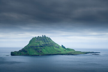 Rocky green island of Tindholmur, in the sea under dark clouds, Faroe Islands.