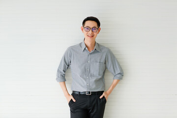 Medium portrait of a formal Asian handsome man with cute smiling, wearing eyeglasses in a gray long sleeve shirt and black pants with hand in pocket posing on white background in the studio