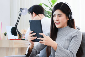 Young Asian black long hair woman staying in living room wearing headphone and holding tablet computer and widely smile to gadget screen while man working blur in blackground.