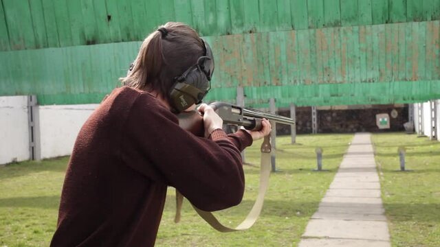 A young man shoots a pump-action firearm at flags, targets. A man in protective glasses and headphones in a dash. A green field with bullet protection outdoor. Sports shooting, weapons hobby. shotgun
