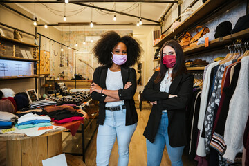 Portrait of two women owners of the dress shop at the entrance to welcome customers during the Coronavirus Covid-19 pandemic wearing protective face masks - Millennial initiate a start-up business - 426636754