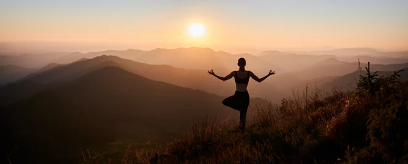 Selbstklebende Fototapete Yogaschule Panoramablick. Rückansicht einer schlanken Frau in schwarzer Sportkleidung, die Yoga-Übungen auf einem Bein in den Bergen macht. Konzept der Yogazeit bei Sonnenuntergang.