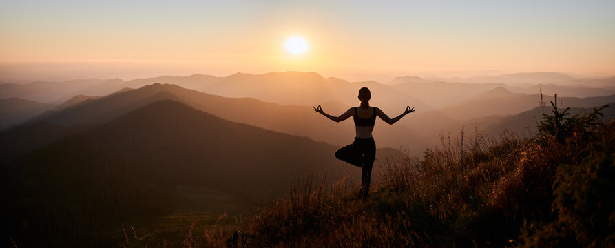 Panoramic view. Back view of slim woman in black sportswear doing yoga exercise on one leg in mountains. Concept of yoga time at sunset.