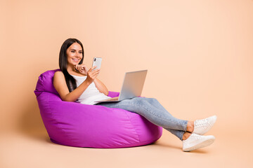 Photo portrait full body view of woman with laptop texting on phone sitting in beanbag chair isolated on pastel beige colored background