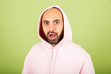 Young bald caucasian man in pink hoodie isolated on green background look to camera shocked with surprise and amazed expression