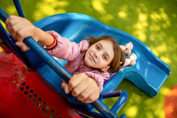 Fototapeta na wymiar cute happy beautiful natural teenage girl in a pink jacket is on a playground with a green special coating hanging on a slide on a sunny day