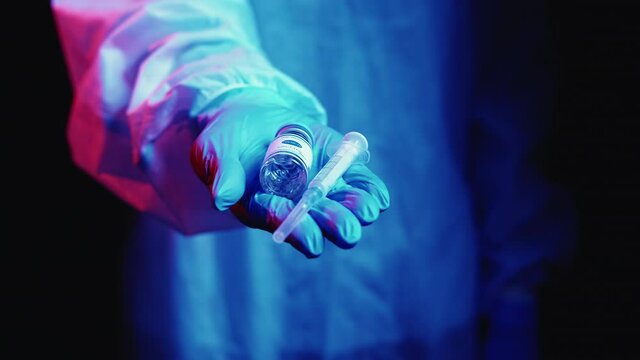 Covid-19 vaccine. Stop pandemic. Global immunization. Doctor in blue ppe protective glove with dose vial syringe in hand in pink neon light on dark blur background.