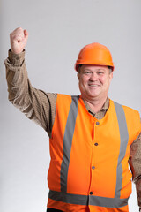 happy construction worker in hard hat with raised hand up on grey studio background, cheerful fat...
