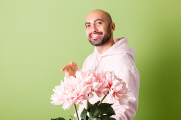 Young bald caucasian man in pink hoodie isolated on green background hold bouquet  of flowers happy smiling look to camera