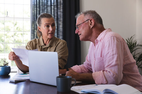 Caucasian senior couple in living room sitting at table using laptop, paying bills and talking
