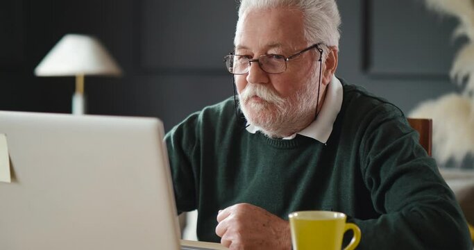 Senior man discusses and explains using a video call on a laptop at home. Grandfather in  having Online Conversation with friend on Laptop