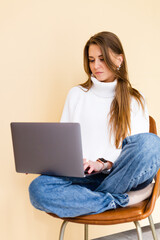 Pretty european woman in casual white sweater isolated on peach background sit on chair with laptop computer on knees businesswoman student freelance