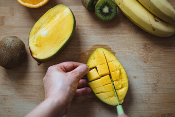 A woman cutting a mango with a green knife on a wooden table surrounded by other fruits (Top view).