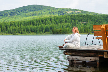 A woman sits on the pier of a blue lake in the mountains and looks into the distance