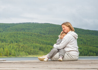 Mom hugs the child sitting next to her on the pier of a blue lake in the mountains and gently looks at the child. Family tourism