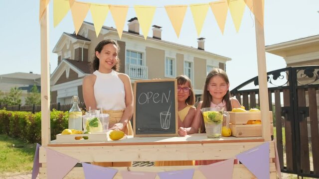 Medium portrait of delighted mixed race woman with two pretty 10-year-old girls smiling to camera standing at diy stand selling lemonade on hot summer day