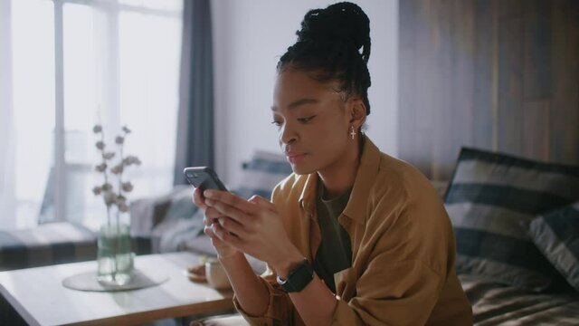 Young afro american woman hold use smart phone sitting at home at sunlight. Watching social media. Portrait, entertainment, smartphone. Slow motion