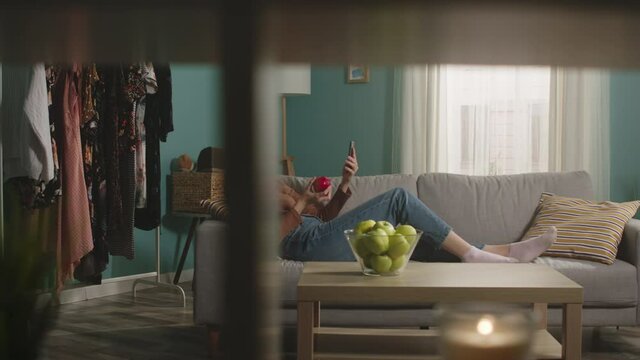 Young woman in blue jeans and brown sweater lies on beige sofa on background of window, biting a red apple and and speaks via video link.
