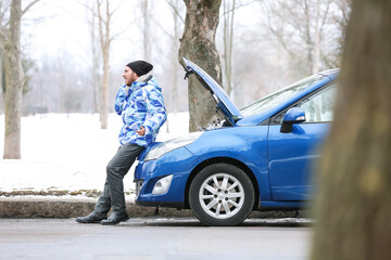 Stressed young man talking by phone near broken car outdoors