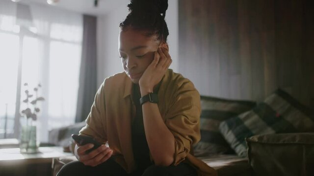 Sad young afro american woman hold use smart phone sitting at home at sunlight. Watching social media. Portrait, smartphone. Slow motion