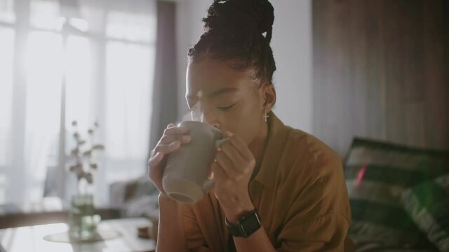 Young beautiful african woman drink cup of coffee or tea sitting at home. Thinking, holding, inspiration, dreaming, cute, positive, relaxation. Close up. Slow motion
