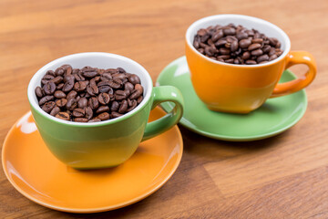 coffee composition, close-up of two cups of coffee one orange and one green with their plates full of coffee beans on a wooden surface, horizontal photo