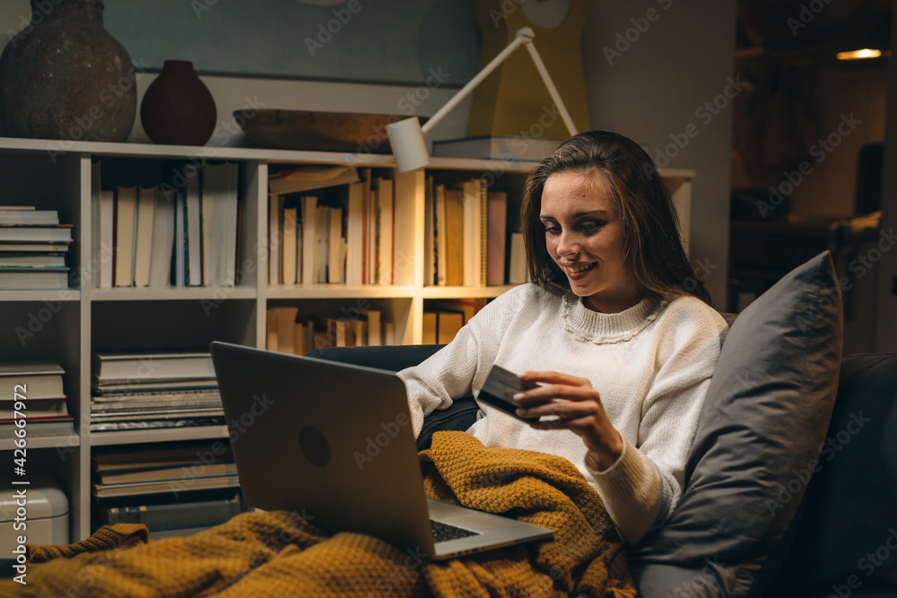 Wall mural woman laying on sofa and shopping online using laptop computer at her home. evening atmosphere